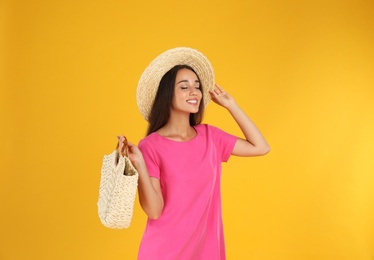 Young woman wearing stylish pink dress with straw bag on yellow background