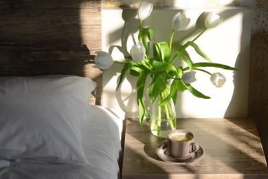 Photo of Beautiful white tulip bouquet and cup of coffee on nightstand in bedroom