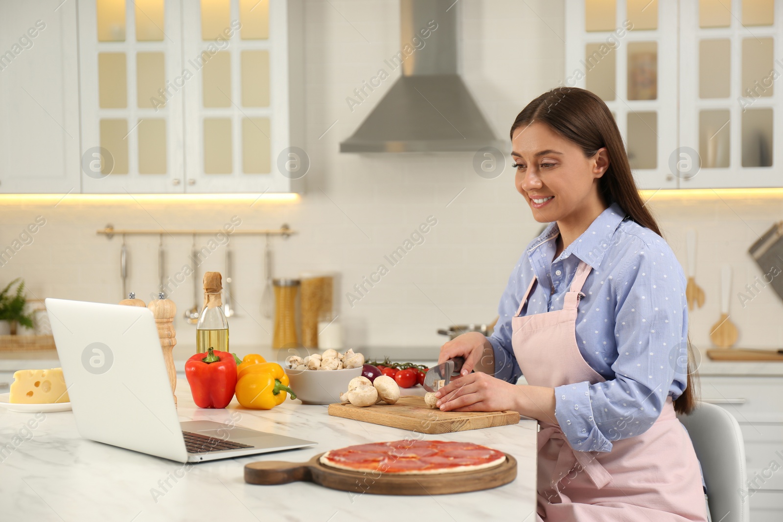 Photo of Happy woman cutting mushroom while watching online cooking course via laptop in kitchen