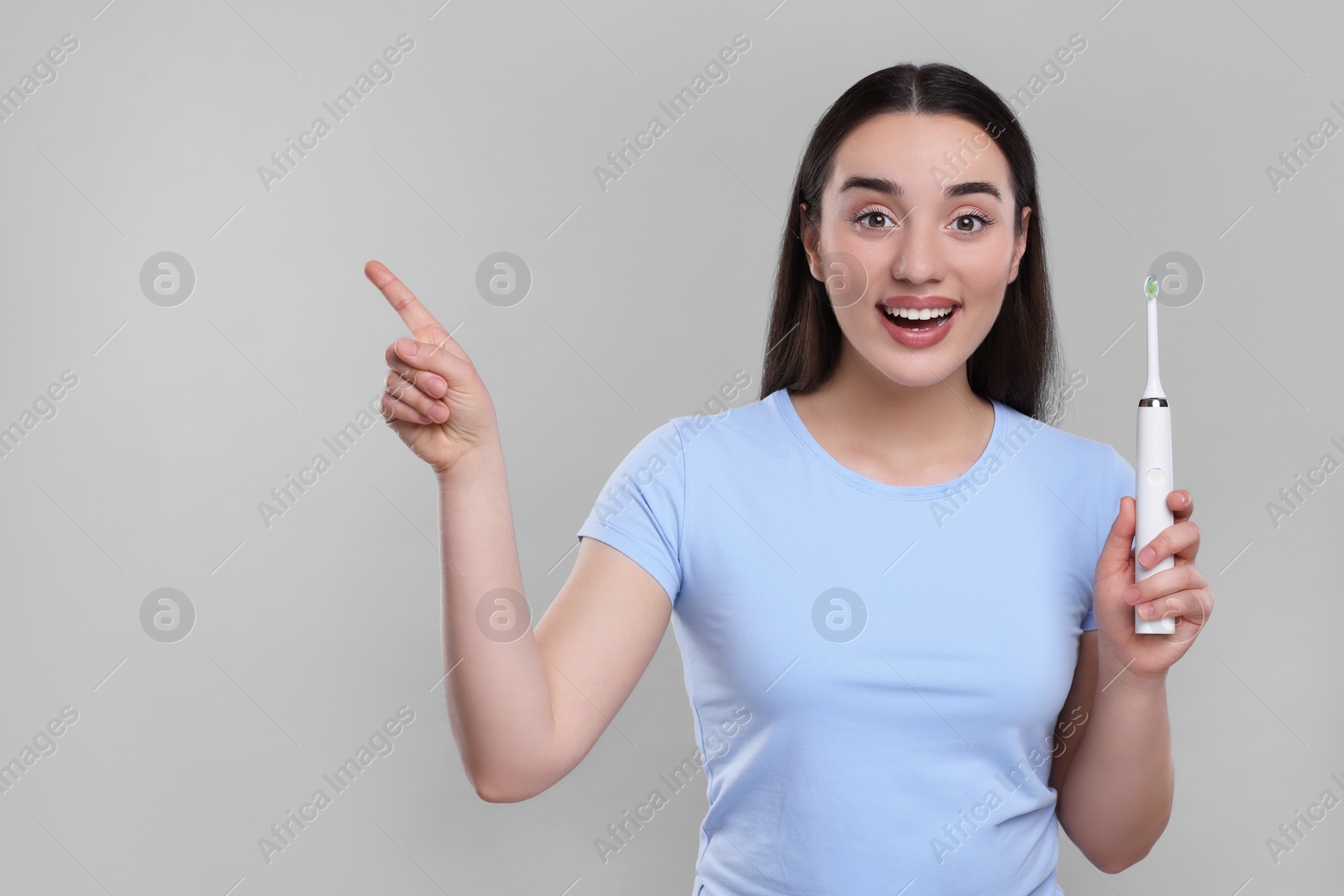Photo of Happy young woman holding electric toothbrush and pointing on light grey background, space for text