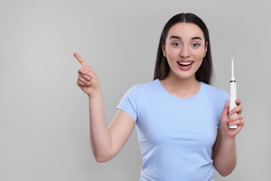 Happy young woman holding electric toothbrush and pointing on light grey background, space for text