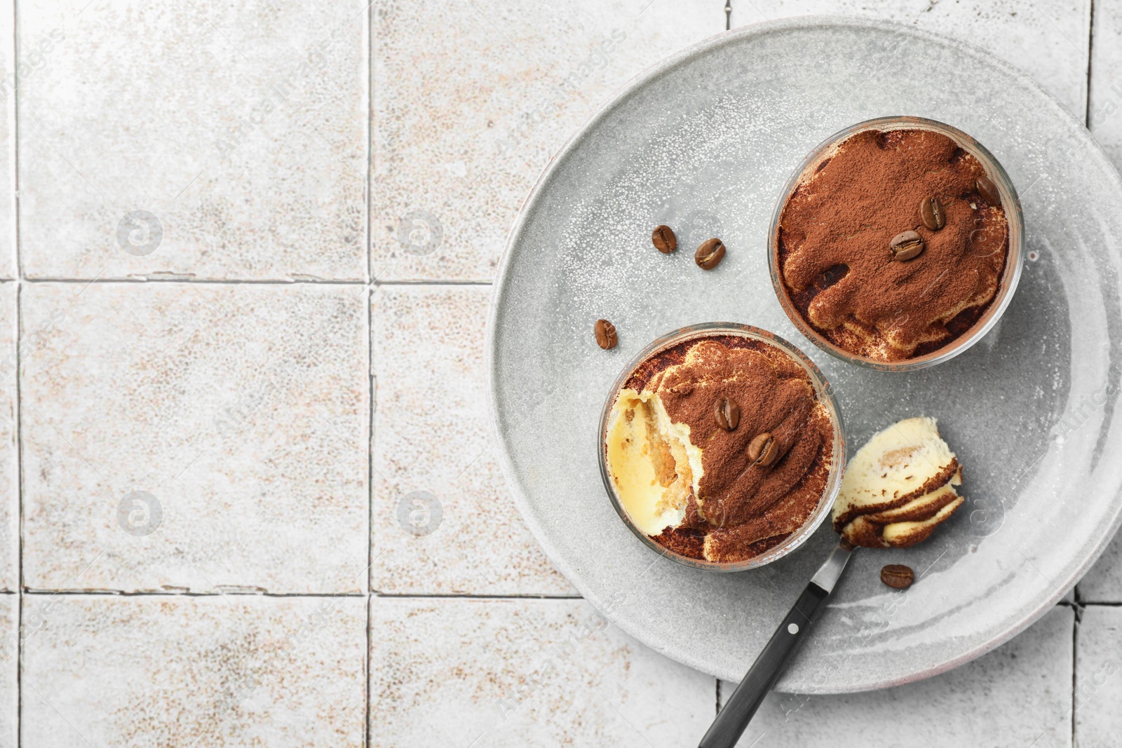 Photo of Delicious tiramisu in glasses, spoon and coffee beans on light tiled table, top view. Space for text