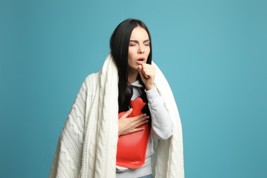 Photo of Ill woman with hot water bottle and knitted blanket coughing on light blue background
