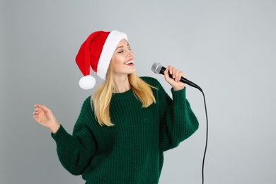 Emotional woman in Santa Claus hat singing with microphone on grey background. Christmas music