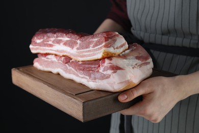 Photo of Woman holding wooden board with pieces of raw pork belly on black background, closeup