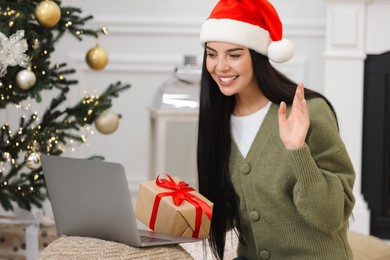 Celebrating Christmas online with exchanged by mail presents. Smiling woman in Santa hat with gift waving hello during video call at home