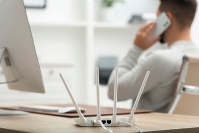 Photo of Man talking on smartphone while working at wooden table indoors, focus on Wi-Fi router