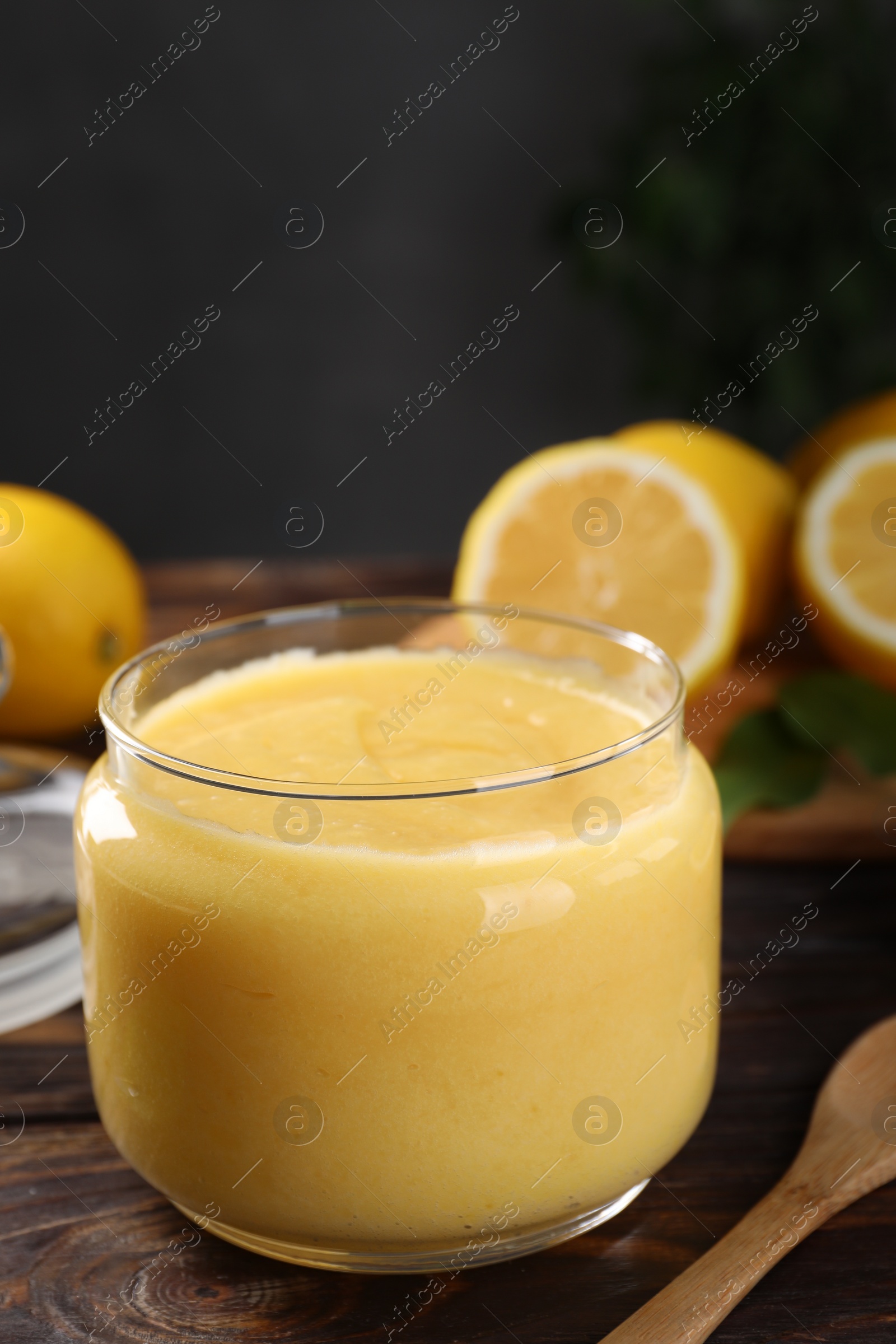 Photo of Delicious lemon curd in glass jar, fresh citrus fruits and spoon on wooden table