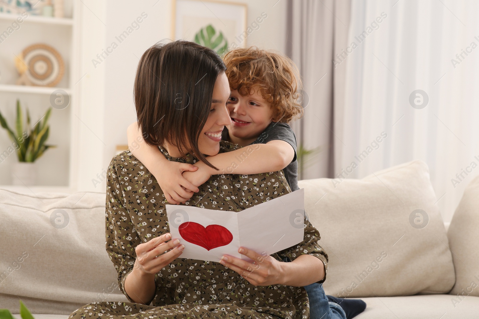 Photo of Little son congratulating his mom with Mother`s day at home. Woman holding handmade greeting card