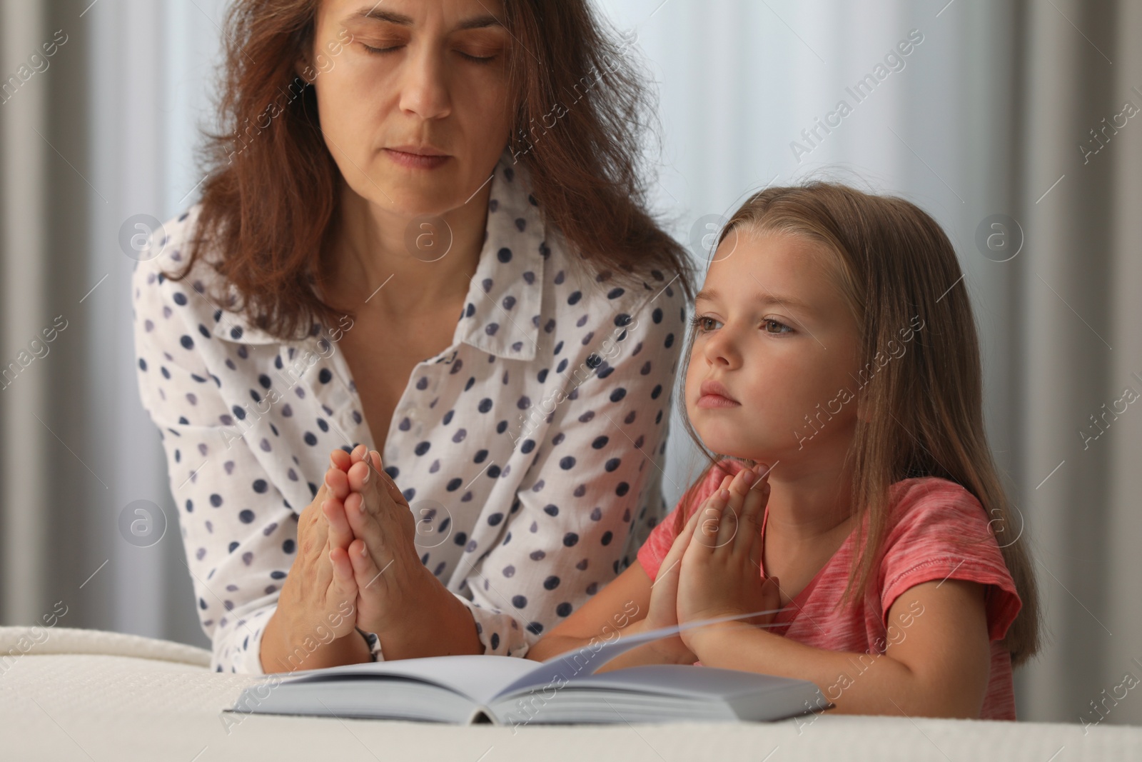 Photo of Mature woman with her little granddaughter praying together over Bible in bedroom