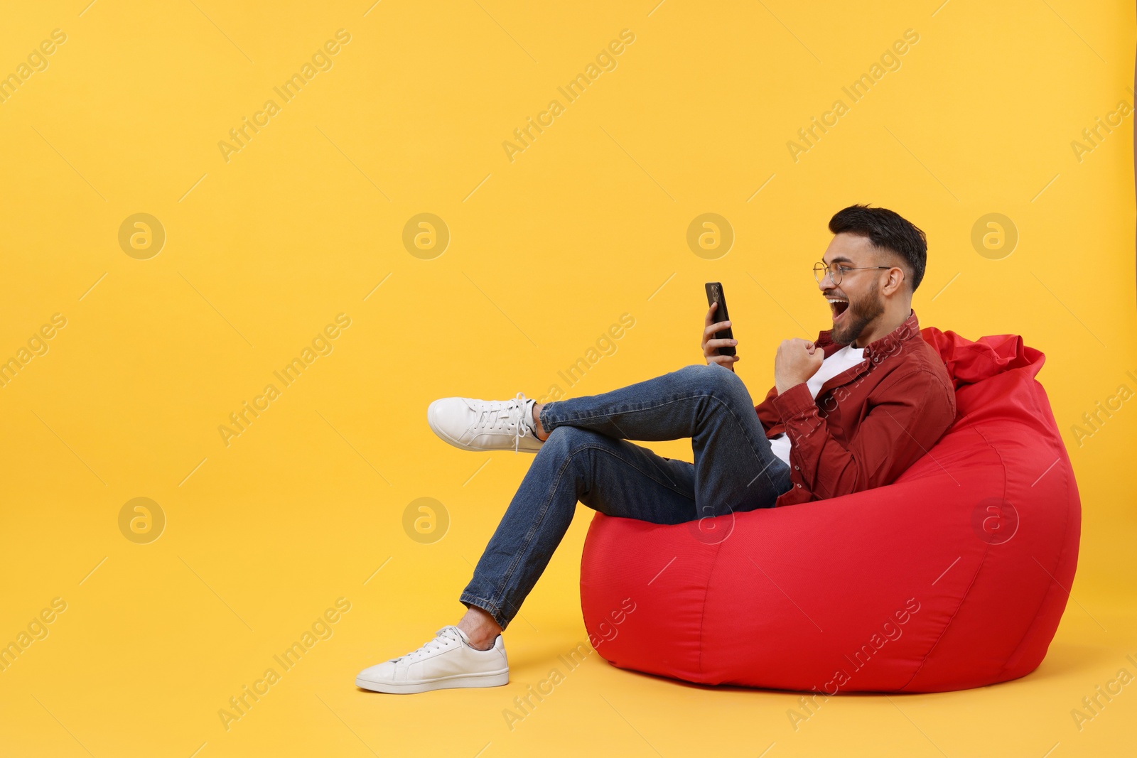 Photo of Happy young man using smartphone on bean bag chair against yellow background, space for text