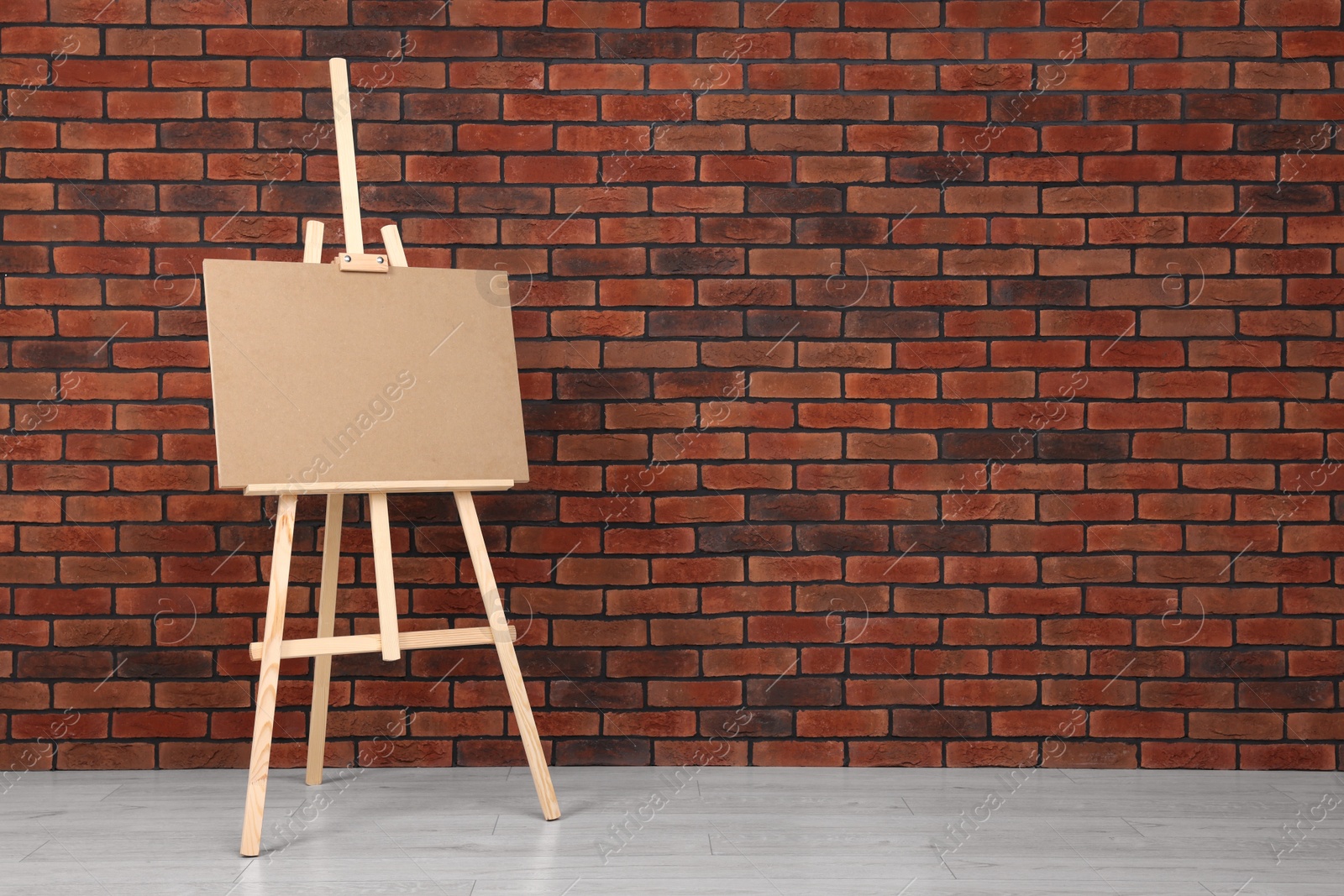 Photo of Wooden easel with blank board near brick wall indoors. Space for text