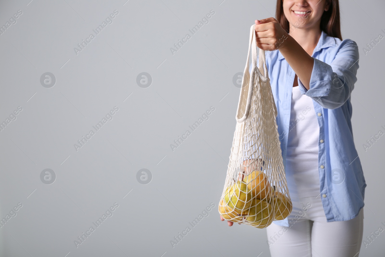 Photo of Woman holding net bag with fresh ripe pears on grey background, closeup. Space for text