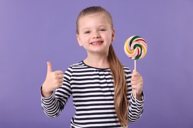 Happy little girl with colorful lollipop swirl showing thumbs up on violet background