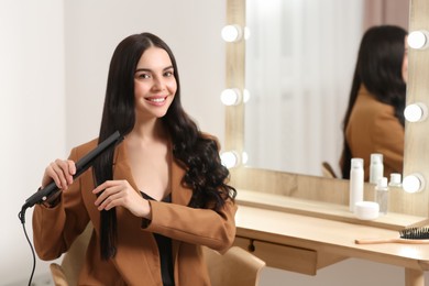 Beautiful happy woman using hair iron near mirror in room. Space for text