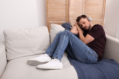 Upset man listening to music through headphones on sofa at home. Loneliness concept