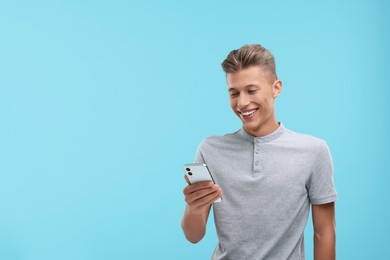 Happy young man sending message via smartphone on light blue background. Space for text