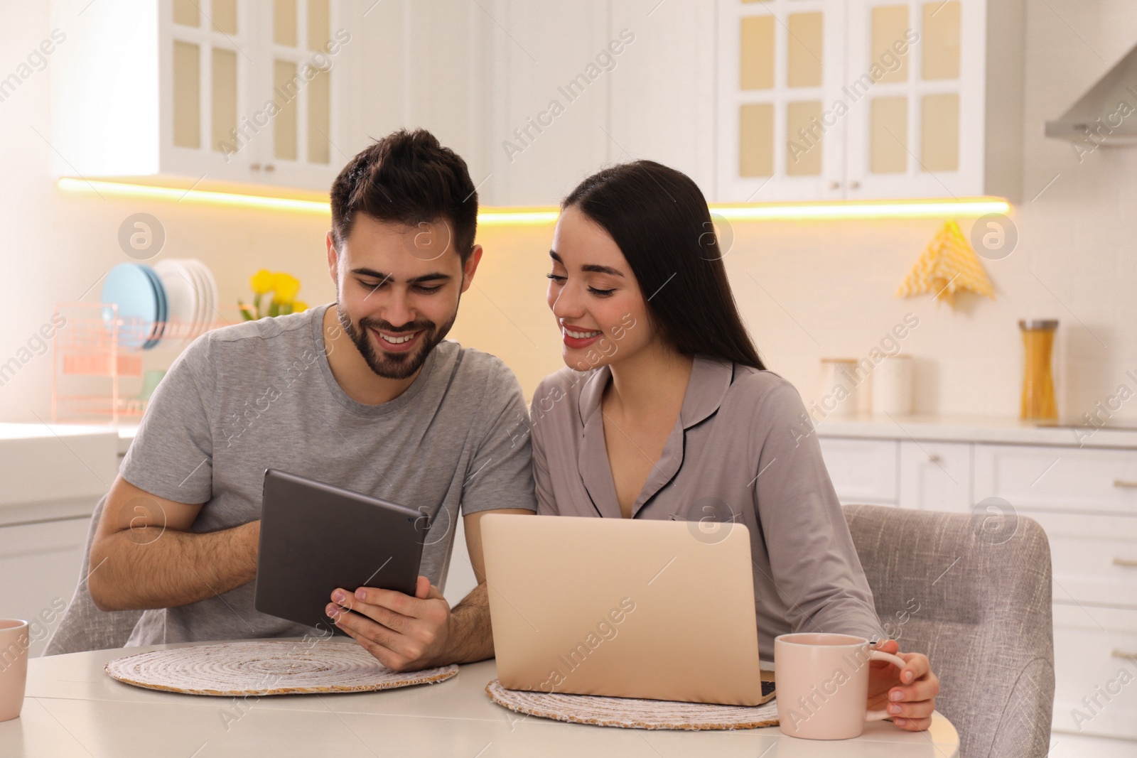 Photo of Happy couple in pajamas using gadgets at kitchen table