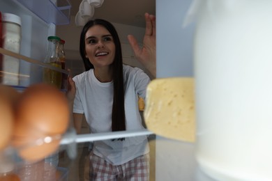 Photo of Young woman near modern refrigerator in kitchen at night, view from inside