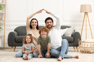 Photo of Family housing concept. Happy woman and her husband forming roof with their hands while sitting with kids on floor at home