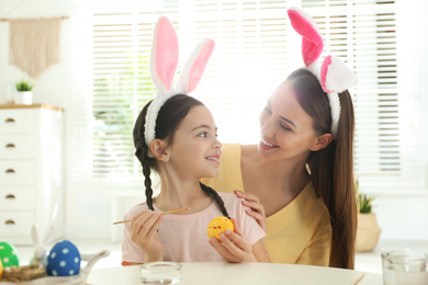 Photo of Happy mother and daughter with bunny ears headbands painting Easter egg at home
