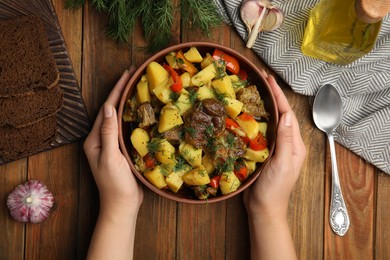 Woman eating delicious dish with potatoes from earthenware at wooden table, top view