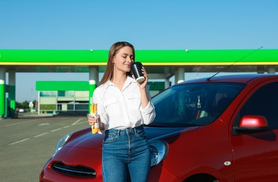 Photo of Beautiful young woman with hot dog drinking coffee near car at gas station
