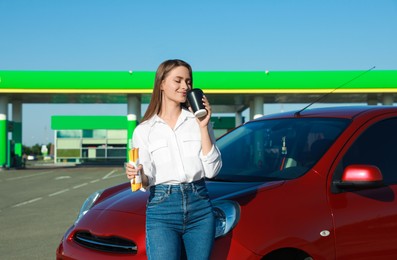 Beautiful young woman with hot dog drinking coffee near car at gas station