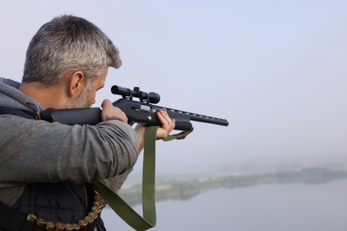 Man aiming with hunting rifle near lake outdoors. Space for text