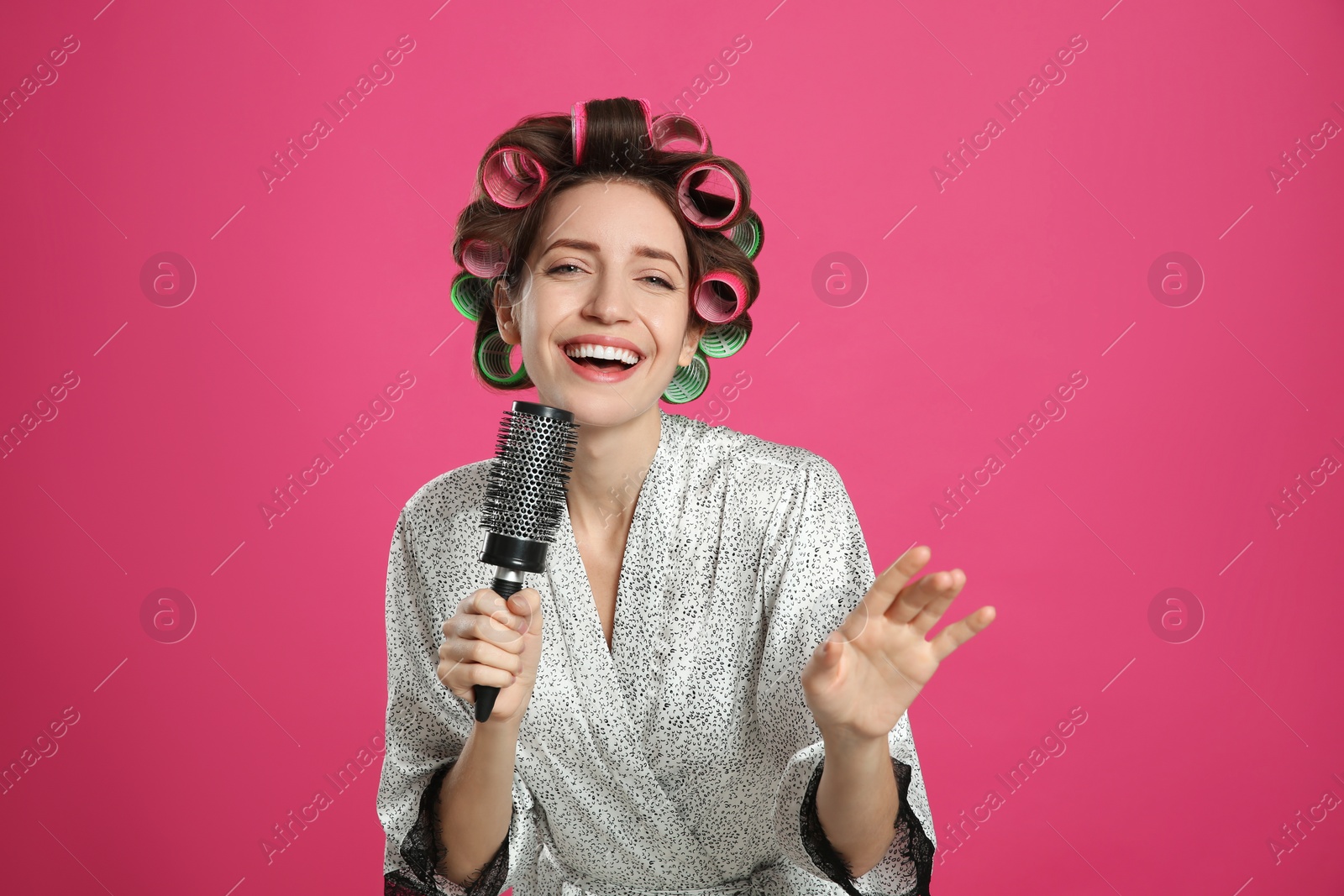 Photo of Beautiful young woman in silk bathrobe with hair curlers singing into hairbrush on pink background