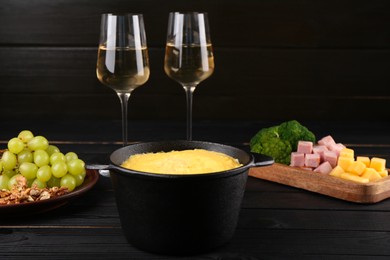 Photo of Fondue pot with melted cheese, glasses of wine and different products on black wooden table