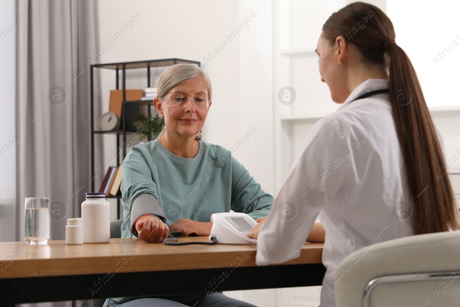 Photo of Young healthcare worker measuring senior woman's blood pressure at wooden table indoors