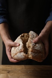 Photo of Man breaking loaf of fresh bread at wooden table, closeup