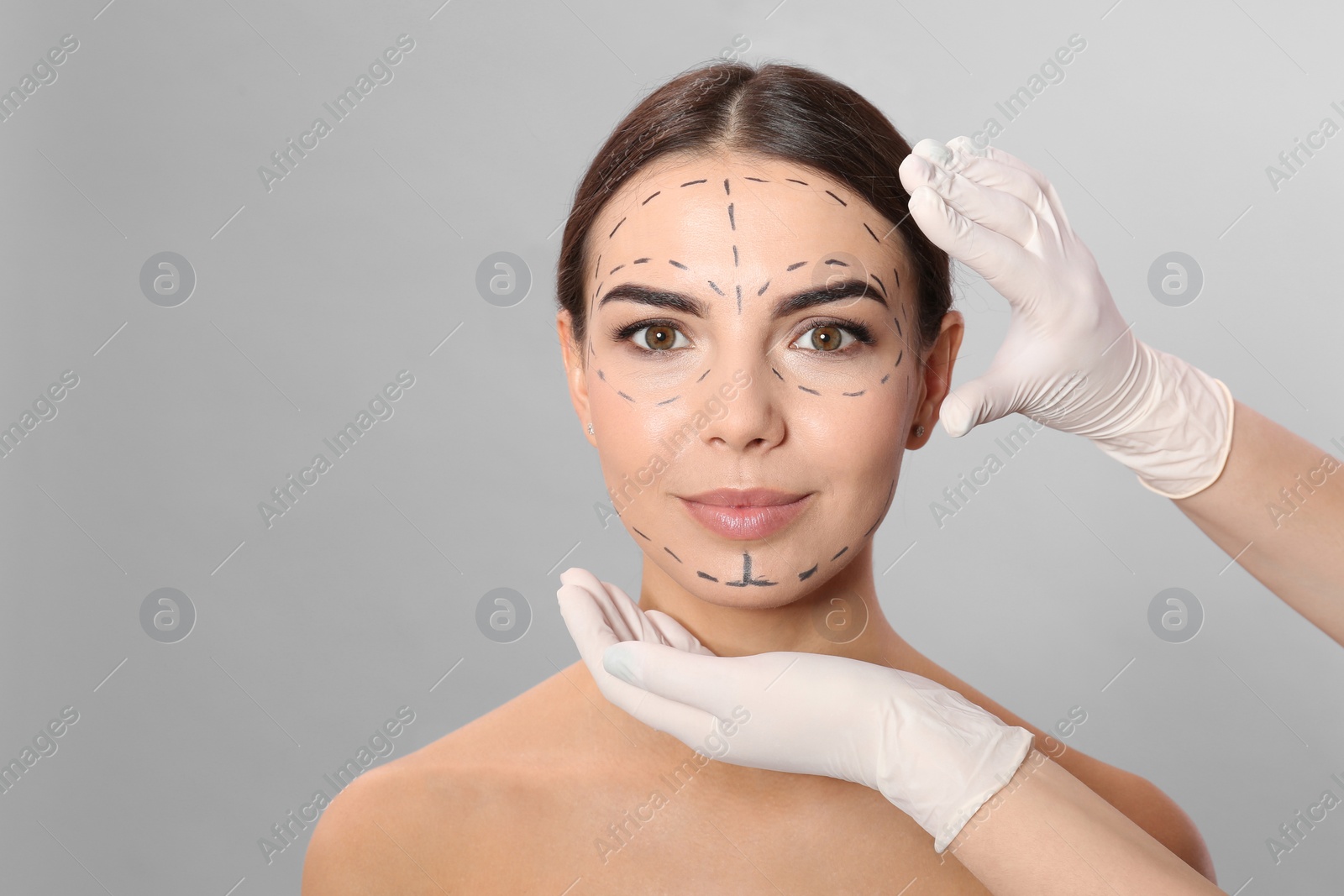 Photo of Doctor examining woman's face with marker lines for plastic surgery operation on grey background