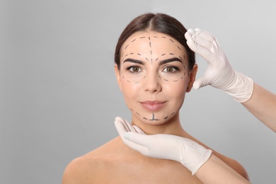 Doctor examining woman's face with marker lines for plastic surgery operation on grey background