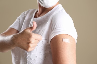 Mature woman in protective mask showing arm with bandage after vaccination and thumb up on beige background, closeup