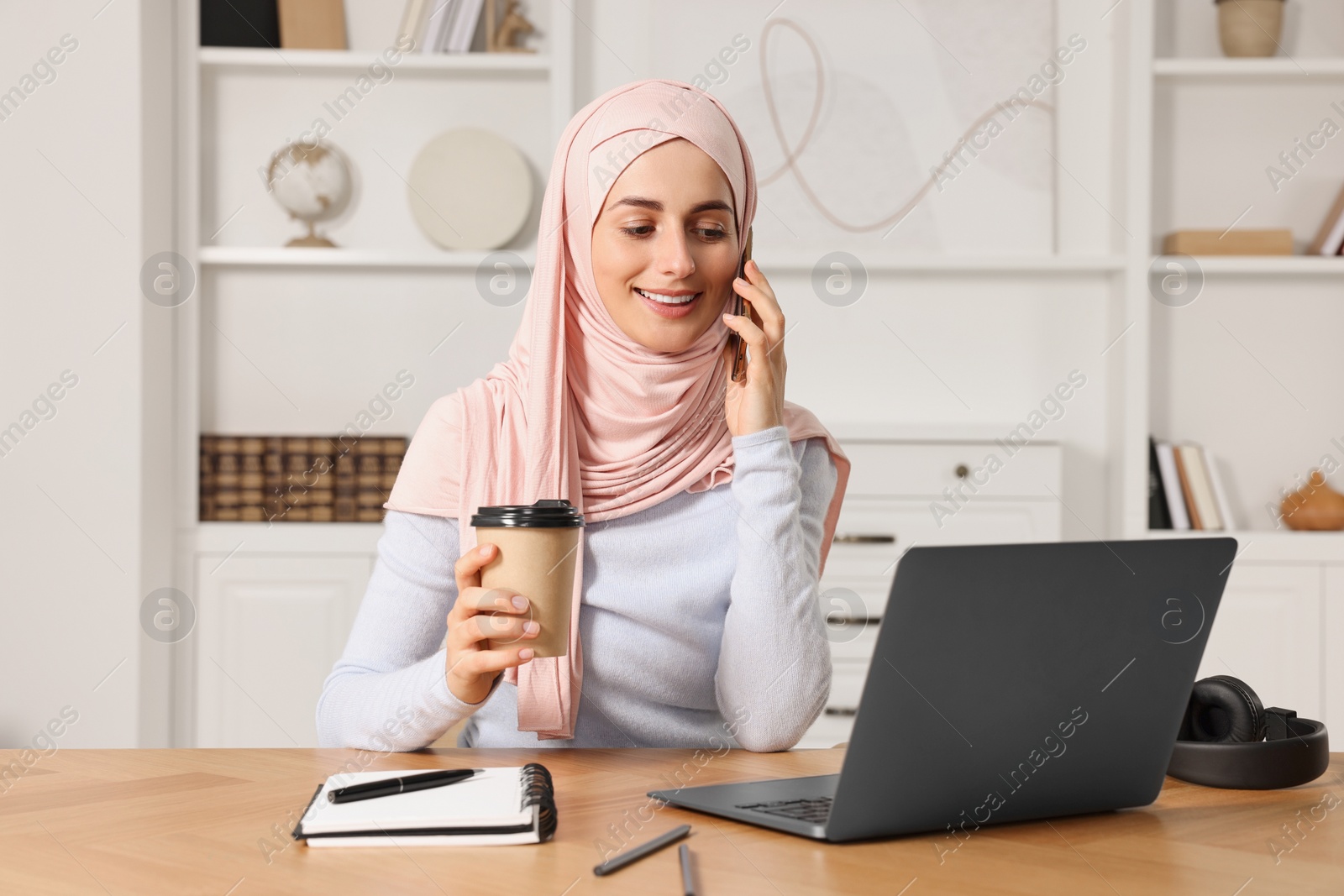 Photo of Muslim woman with cup of coffee talking on smartphone near laptop at wooden table in room