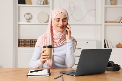 Muslim woman with cup of coffee talking on smartphone near laptop at wooden table in room