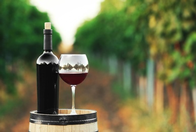 Photo of Bottle and glass of red wine on wooden barrel in vineyard