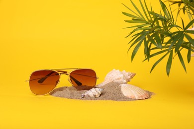Photo of Stylish sunglasses, seashells, sand and palm leaves on yellow background