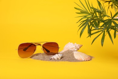 Stylish sunglasses, seashells, sand and palm leaves on yellow background