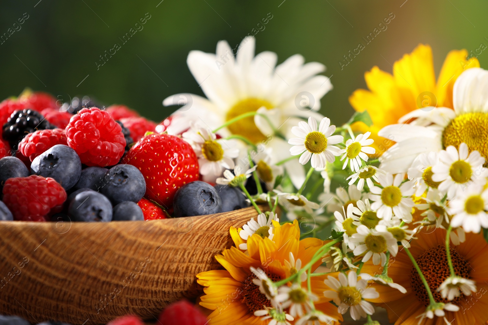 Photo of Bowl with different fresh ripe berries and beautiful flowers on table outdoors, closeup