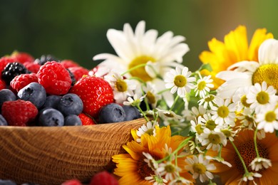 Photo of Bowl with different fresh ripe berries and beautiful flowers on table outdoors, closeup