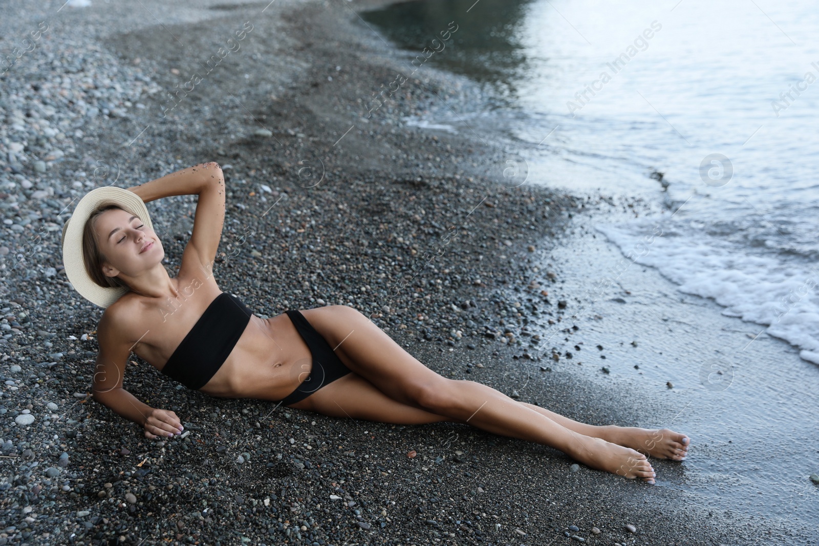 Photo of Beautiful young woman in stylish bikini and straw hat lying on pebble seashore