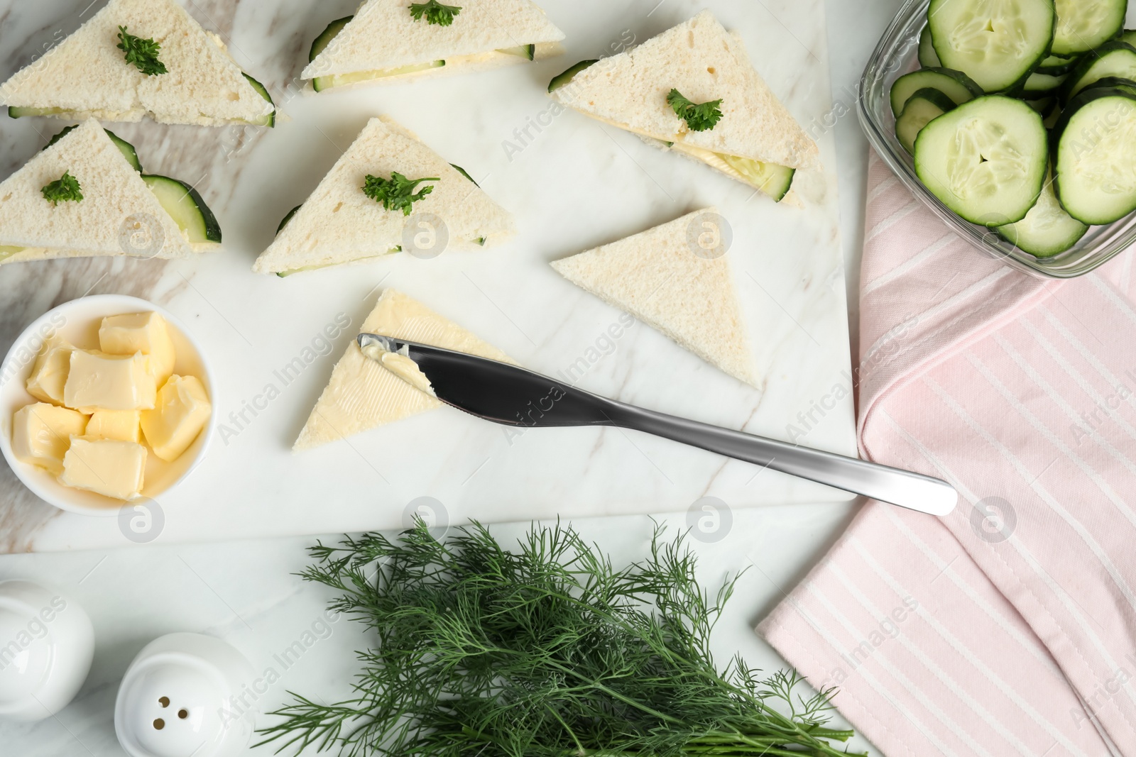 Photo of Spreading butter on tasty sandwiches with cucumber and parsley on white marble table, flat lay