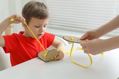 Mother teaching son to tie shoe laces using training cardboard template at white table, closeup