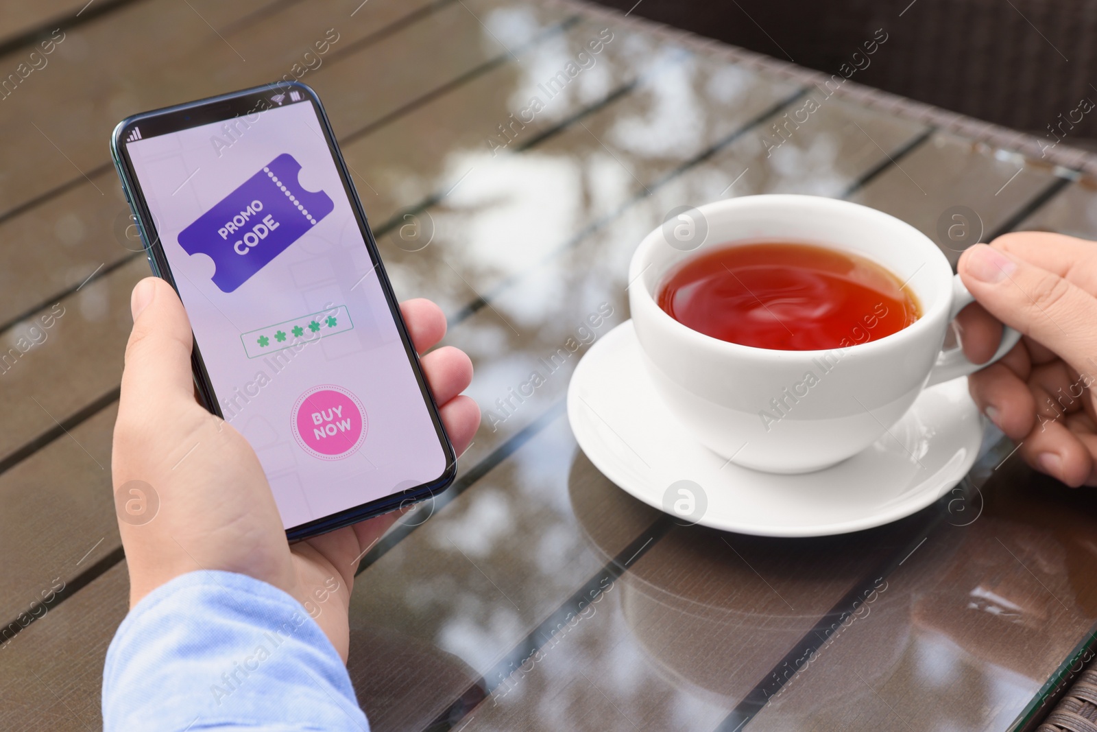 Photo of Man holding smartphone with promo code and cup of tea at wooden table outdoors, closeup