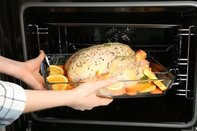 Woman putting raw chicken with orange slices into oven, closeup