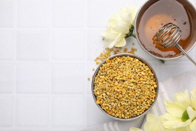 Fresh bee pollen granules, flowers, honey and dipper on white tiled table, flat lay. Space for text