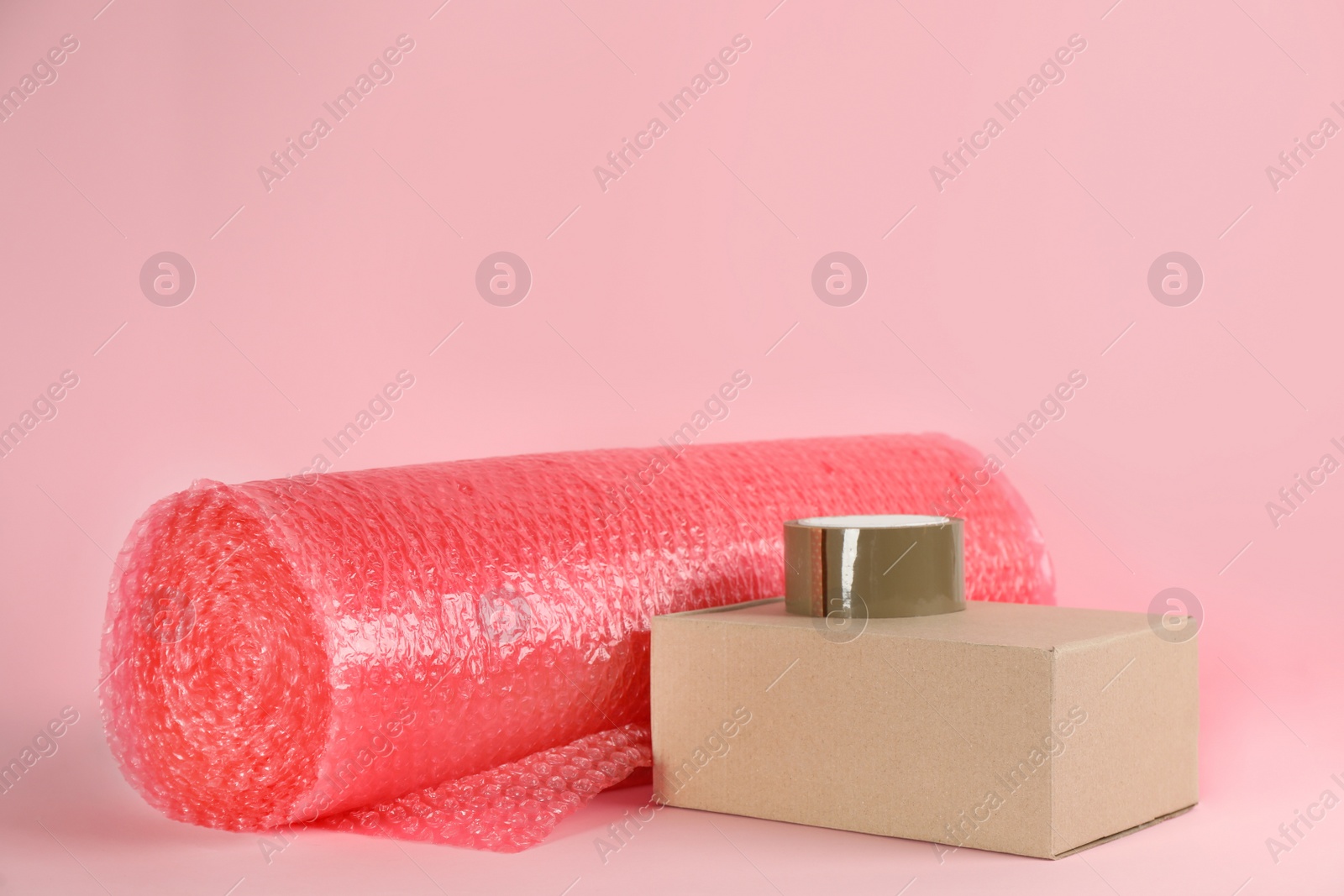 Photo of Bubble wrap roll, adhesive tape and cardboard box on pink background, space for text